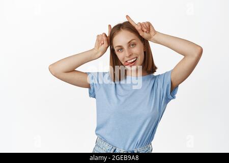 Giovane donna bionda spensierata mostra lingua, corna toro diavolo gesto sulla testa, sorridente spensierato, divertirsi, essere giocoso e sciocco, in piedi in t-shirt Foto Stock