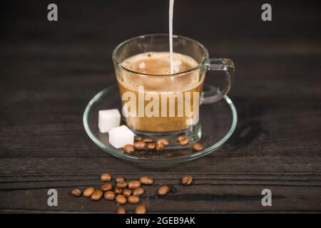 Una tazza di vetro, su un piattino, riempita con caffè estratto. Un flusso di latte bianco si versa nel caffè dall'alto. Foto Stock