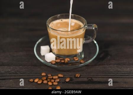 Una tazza di vetro, su un piattino, riempita con caffè estratto. Un flusso di latte bianco si versa nel caffè dall'alto. Foto Stock