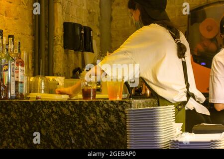 Una barista femminile che indossa una maschera medica prepara cocktail. Cafe operaio, bistrot. Foto Stock