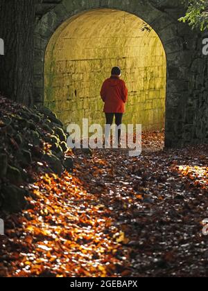 Donna lone con cappello e cappotto rosso che cammina nel tunnel arcuato di pietra pieno di luce verde a Cumbria, Inghilterra, Regno Unito Foto Stock