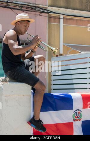 Uomo latino che suona una tromba seduto su un muro con la bandiera della Repubblica Dominicana: Fuoco selettivo. Foto Stock