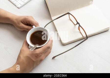 su una scrivania una persona che tiene una tazza di caffè, con un notebook con fogli vuoti aperti e bicchieri moderni, stile di vita sul luogo di lavoro, studio Foto Stock