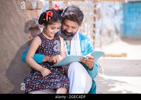 Felice padre indiano rurale aiutando sua figlia negli studi, adorabile bambina del villaggio bambino tenendo notebook studiare fuori casa. L'uomo insegna e. Foto Stock