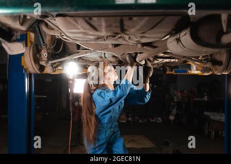 Giovane bella donna meccanica auto vestita con occhiali protettivi e tute da lavoro fa un controllo del telaio della vettura. Spazio di copia. Foto Stock