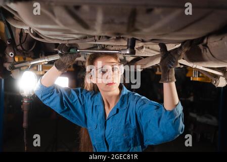 Giovane bella donna meccanica auto è vestita in occhiali di sicurezza e tute da lavoro gira un dado per le chiavi. Spazio di copia. Foto Stock