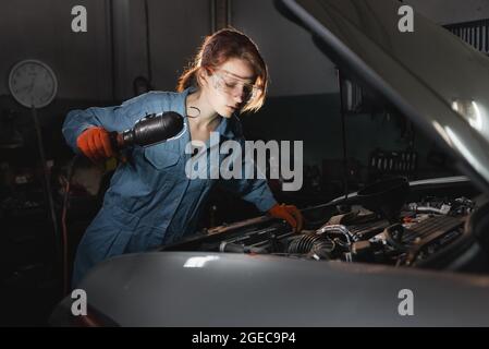 Una meccanica donna effettua un'ispezione di un motore di auto in una stazione di servizio vestita con tute da lavoro in uniforme. Foto Stock