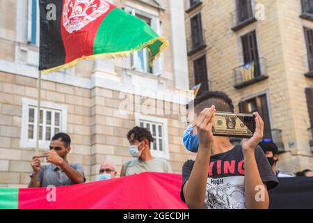 Barcellona, Catalogna, Spagna. 18 agosto 2021. Un ragazzo con un cellulare con un caso del dollaro è visto di fronte a manifestanti con una bandiera afgana.circa cinquecento persone hanno dimostrato a Barcellona in solidarietà con le ragazze e le donne dell'Afghanistan e in difesa dei loro diritti sotto lo slogan ''Afghanistan: Per una vita dignitosa e liberta' (Credit Image: © Thiago Prudencio/DAX via ZUMA Press Wire) Foto Stock