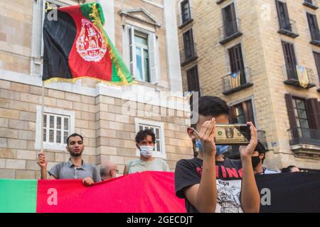 Barcellona, Catalogna, Spagna. 18 agosto 2021. Un ragazzo che guarda un cellulare con un caso di un dollaro è visto di fronte a manifestanti con una bandiera afgana.circa cinquecento persone hanno dimostrato a Barcellona in solidarietà con le ragazze e le donne dell'Afghanistan e in difesa dei loro diritti sotto lo slogan ''Afghanistan: Per una vita dignitosa e liberta' (Credit Image: © Thiago Prudencio/DAX via ZUMA Press Wire) Foto Stock