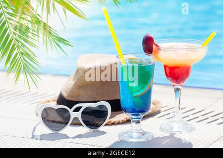 Due colorati bicchieri da cocktail, occhiali da sole e cappello al bordo della piscina Foto Stock