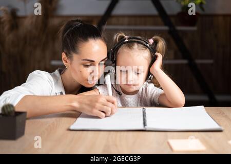 Felice giovane mamma caucasica e la piccola figlia di scrittura a mano in taccuino preparare la scuola a casa compito insieme. Una madre premurosa aiuta sua figlia. Distanza e. Foto Stock