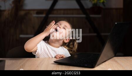 Una ragazza prescolare la sera si siede a un tavolo con un computer portatile e sbadiglii. Foto Stock