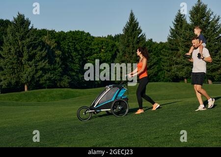 Giornata in famiglia. Una giovane famiglia felice con un bambino passa il tempo insieme mentre cammina con la carrozza del bambino su un prato verde in un parco della città in un fine settimana Foto Stock