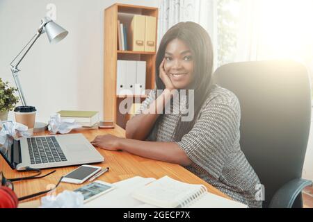Giovane donna d'affari sorridente seduto alla scrivania dell'ufficio con un portatile aperto e un pianificatore davanti a lei e guardando la fotocamera Foto Stock