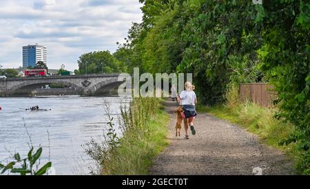 Kew , Londra UK 19 agosto 2021 - UN corridore e il suo cane gode di una miscela di sole e nuvole sulla riva del fiume Tamigi a Kew a Londra con le previsioni del tempo per migliorare all'inizio della prossima settimana in tutto il Regno Unito. : Credit Simon Dack / Alamy Live News Foto Stock