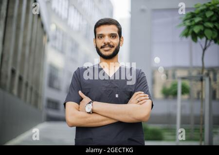 Primo piano ritratto di giovane professionista medico arabo maschio, indossando abiti grigio scrub medico, sorridente e guardando la macchina fotografica, in piedi all'aperto a fro Foto Stock