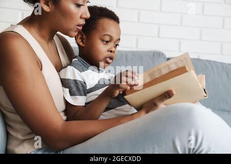 Libro di lettura di madre e figlio neri mentre si siede sul divano a casa Foto Stock