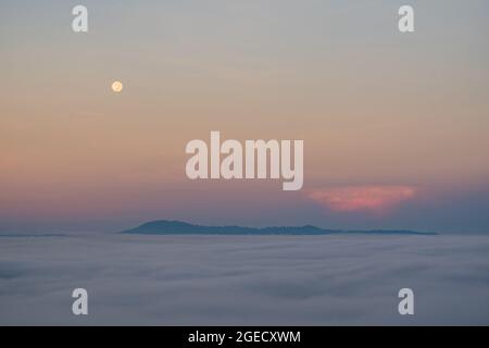 Luna piena che cade e la nebbia al mattino. Il colore arancione del cielo riflette i raggi del sole. Foto Stock