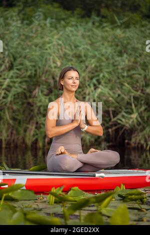 Una giovane caucasica bella ragazza snella che fa yoga, pilates su tavola di pagaia, SUP solo in estate mattina. Concetto di vita attiva, sport, attività per il tempo libero Foto Stock