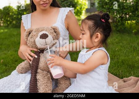 ragazza asiatica del toddler che tiene la tazza di plastica con il mungshake vicino l'orso del toddy nelle mani della madre Foto Stock