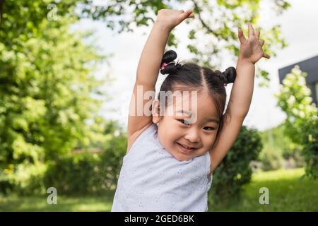 giocoso bambino asiatico in piedi con le mani sollevate e sorridente all'esterno Foto Stock