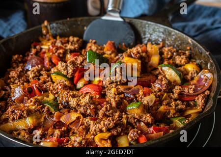 Mescolare il trito di manzo fritto con le verdure. Piatto mediterraneo con zucchine, peperoni, cipolle rosse, aglio ed erbe serviti in una padella in ghisa Foto Stock