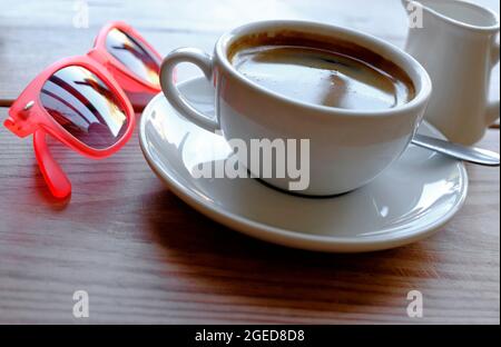 tazza di caffè americano sul tavolino da caffè e occhiali da sole rossi Foto Stock