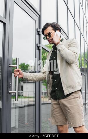 elegante giovane uomo in occhiali da sole che parla sul telefono cellulare mentre si entra in costruzione Foto Stock