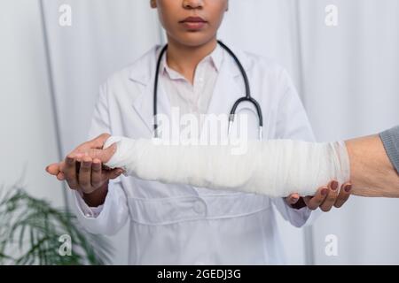 Vista ritagliata di un medico afroamericano sfocato che tocca la mano di una donna anziana nel bendaggio di intonaco Foto Stock