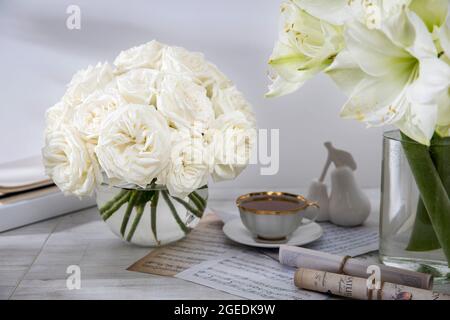 Londra, Regno Unito -20 marzo 2021, un bouquet di rose bianche in un vaso di vetro rotondo su un tavolo con una tazza di tè e un libro. Spazio di copia. Foto Stock