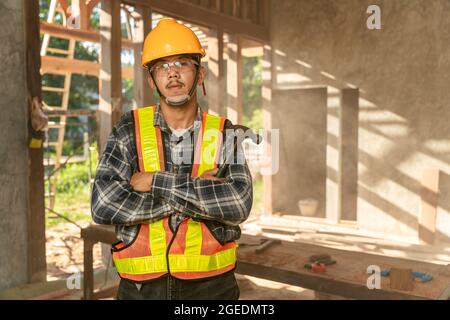 Artigiano asiatico con tenere un martello in mano in piedi in officina spaziosa e guardare la macchina fotografica sul cantiere Foto Stock