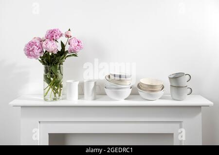 Bouquet di peonie rosa, una pila di tazze, piatti, ciotole, cassettiera bianca all'interno della cucina Foto Stock