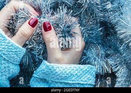 Le mani femminili in un maglione a maglia blu con una bella manicure lucida - borgogna, rosso scuro, chiodi color ciliegia sullo sfondo di latta di Natale d'argento Foto Stock