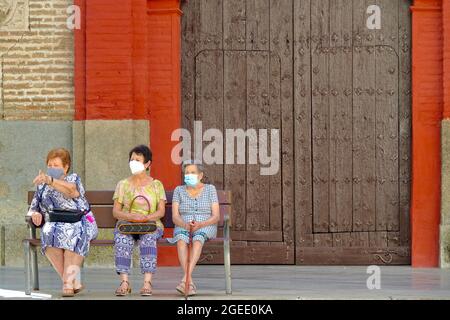 Güéjar-Sierra (Granada), Spagna; agosto-19, 2021: Tre donne di età diversa, indossando maschere, seduti su una panchina accanto ad un cancello chiuso di legno Foto Stock