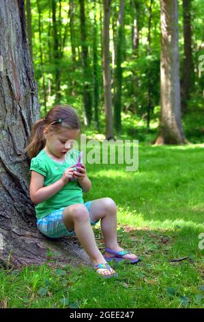 La bambina finge di chiamare un amico sul suo telefono di gioco. Indossa pantaloncini a plaid e una parte superiore verde. Lei è seduta accanto ad un albero sul gr Foto Stock