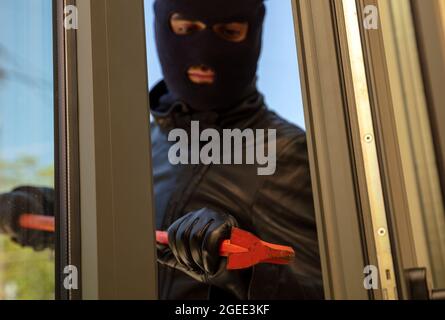 Housebreak con crowbar. Burglar in balaclava che rompe una finestra di vetro. Ladro mascherato che entra in una casa per rubare di giorno. Foto Stock