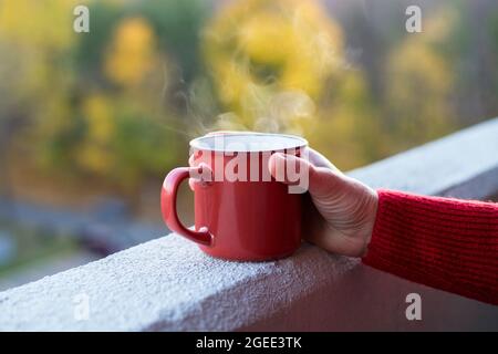 Tazza di bevanda calda in mano della donna si erge sul bordo del balcone in un appartamento alto o edificio di uffici, con alberi sfocati e tramonto sullo sfondo. Foto Stock