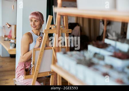 Positivo giovane femmina modello con capelli rosa siede a cavalletto in legno con tela Foto Stock