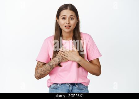 La giovane donna sorpresa sembra lusingata e grata, tiene le mani sul petto e sorride, si erge su sfondo bianco Foto Stock