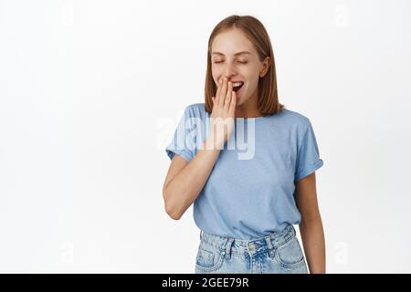 Immagine di una ragazza bionda soddisfatta, sbadigliata di piacere, ha avuto una buona notte di sonno, un grande pisolino, occhi vicini e sorriso durante l'alba, coprire la bocca con la mano Foto Stock