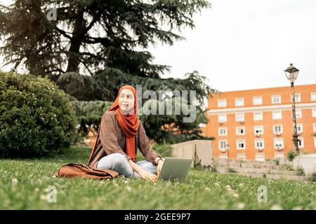 Foto di stock di donna musulmana che usa il computer che si siede sull'erba. Indossa un hiyab Foto Stock