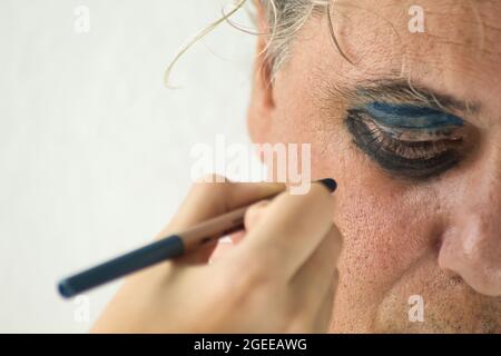 mano di una ragazza che dipinge gli occhi di un uomo maturo serio con cosmetico su sfondo bianco Foto Stock