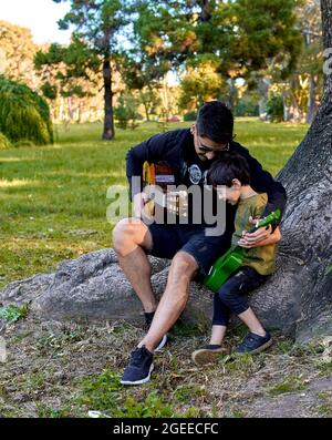 Padre latino abbraccia suo figlio godendo di suonare la chitarra in foresta seduta in un albero. Verticale Foto Stock