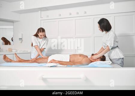 Le donne chiropratica in uniformi lavorano con pazienti maschi maturi sdraiati sul divano in un salone termale Foto Stock