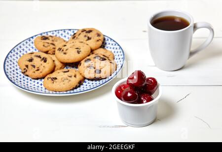 Tazza bianca con caffè, biscotti tradizionali e ciliegie dolci in una ciotola su sfondo bianco di legno, primo piano. Colazione gustosa, colazione servita ogni mattina Foto Stock