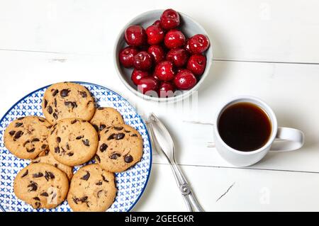 Tazza bianca con caffè, biscotti tradizionali sul piatto e ciliegie dolci in una ciotola di ceramica su sfondo bianco di legno, primo piano. Colazione gustosa, morni Foto Stock