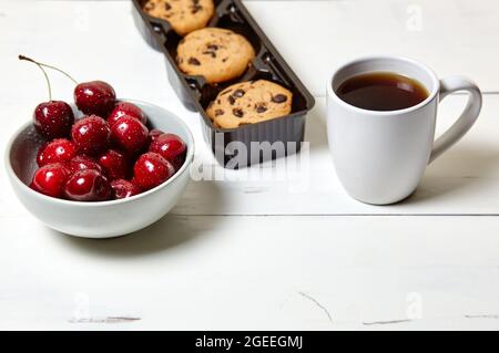 Tazza bianca con caffè, biscotti tradizionali e ciliegie dolci in una ciotola di ceramica su fondo di legno bianco, primo piano. Gustosa colazione, mattina routin Foto Stock