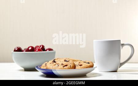 Tazza bianca con caffè, biscotti tradizionali e ciliegie dolci in una ciotola di ceramica su fondo di legno bianco, primo piano. Gustosa colazione, mattina routin Foto Stock