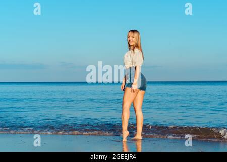 Fitness donna sorridente in cima bianca e jeans corti in posa su sfondo blu mare e cielo durante una passeggiata sulla spiaggia di mare. Vacanze estive Foto Stock