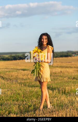 Ritratto di una bella donna sorridente dai capelli scuri con bouquet di girasoli sul campo raccolto Foto Stock